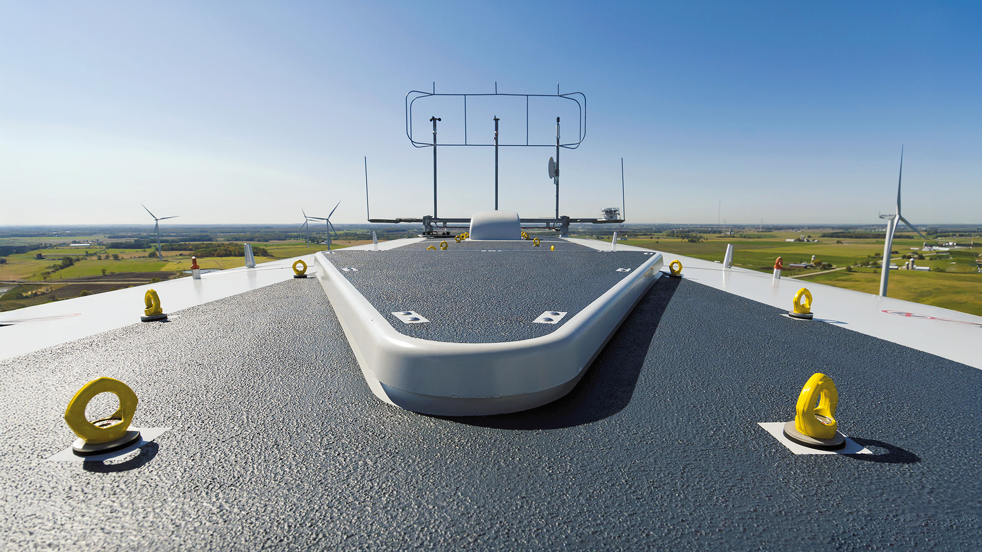 Blick über das Dach einer Windkraftanlage mit mehreren gelben RUD-Personenanschlagpunkten zur Absturzsicherung. Im Hintergrund sind weitere Windräder in einer weiten Landschaft zu sehen, bei klarem Himmel und guter Sicht.